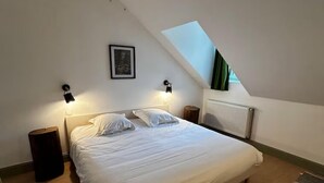 A bedroom with a sloped ceiling, a bed with white bedding, two bedside lamps, a framed picture on the wall, and a radiator. - Gîte La Garenne (La Ferté-Saint-Aubin)