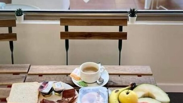 A breakfast setup on a wooden table with a white decorative piece, a cup of coffee, toast, ham, eggs, fruit, and a glass of orange juice. - ALBERGUE AS POZAS TERMAIS (Caldas de Reis)