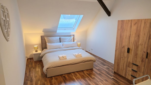A bedroom with a skylight, wooden flooring, a bed with pillows and a blanket, bedside lamps, and a wooden wardrobe. - Superior apartment with private balcony & parking space (Chemnitz)