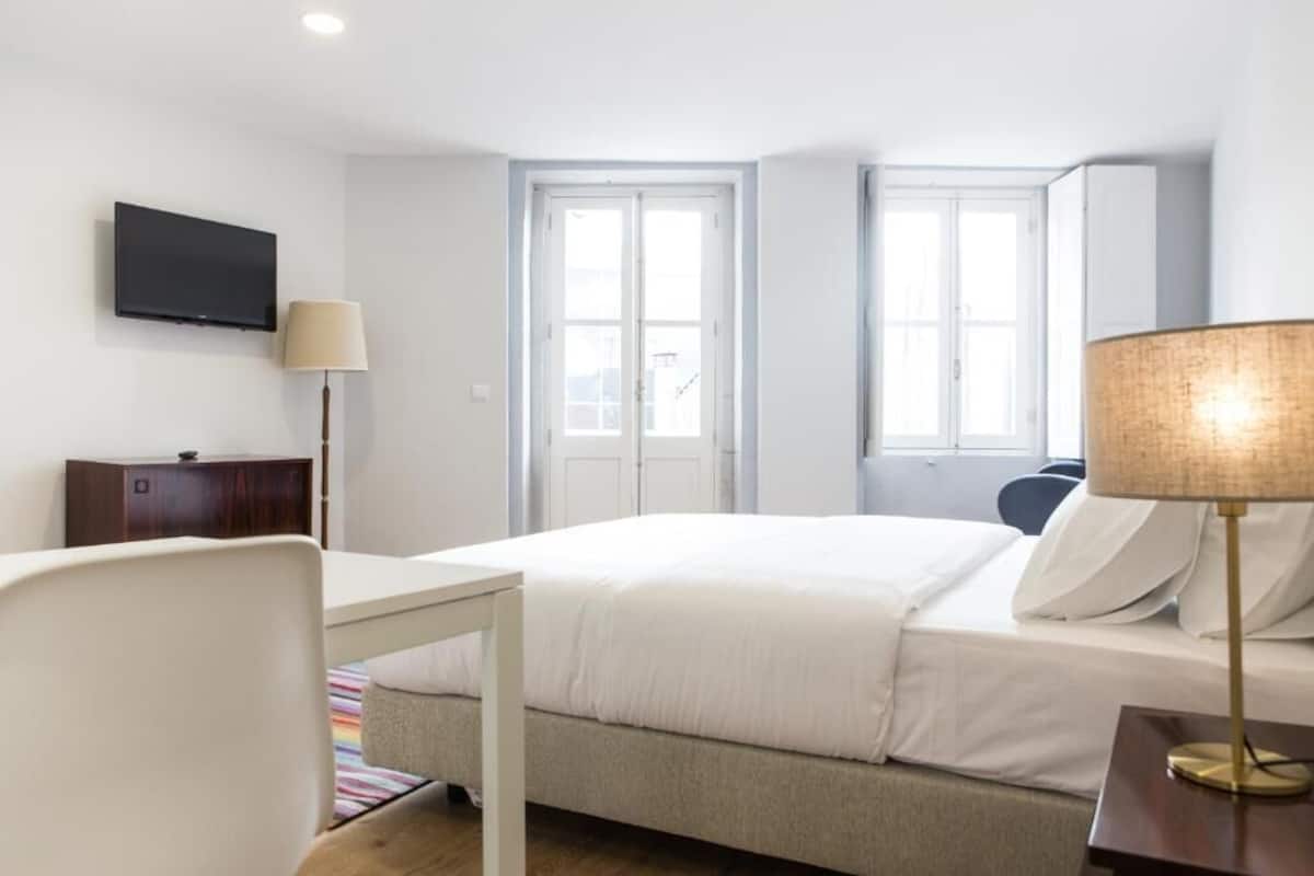 A hotel room with a bed, a desk, a chair, a TV, and two windows.