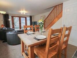 Un comedor con una mesa de madera, cuatro sillas y un florero con flores. En el fondo, una escalera conduce a una puerta.
