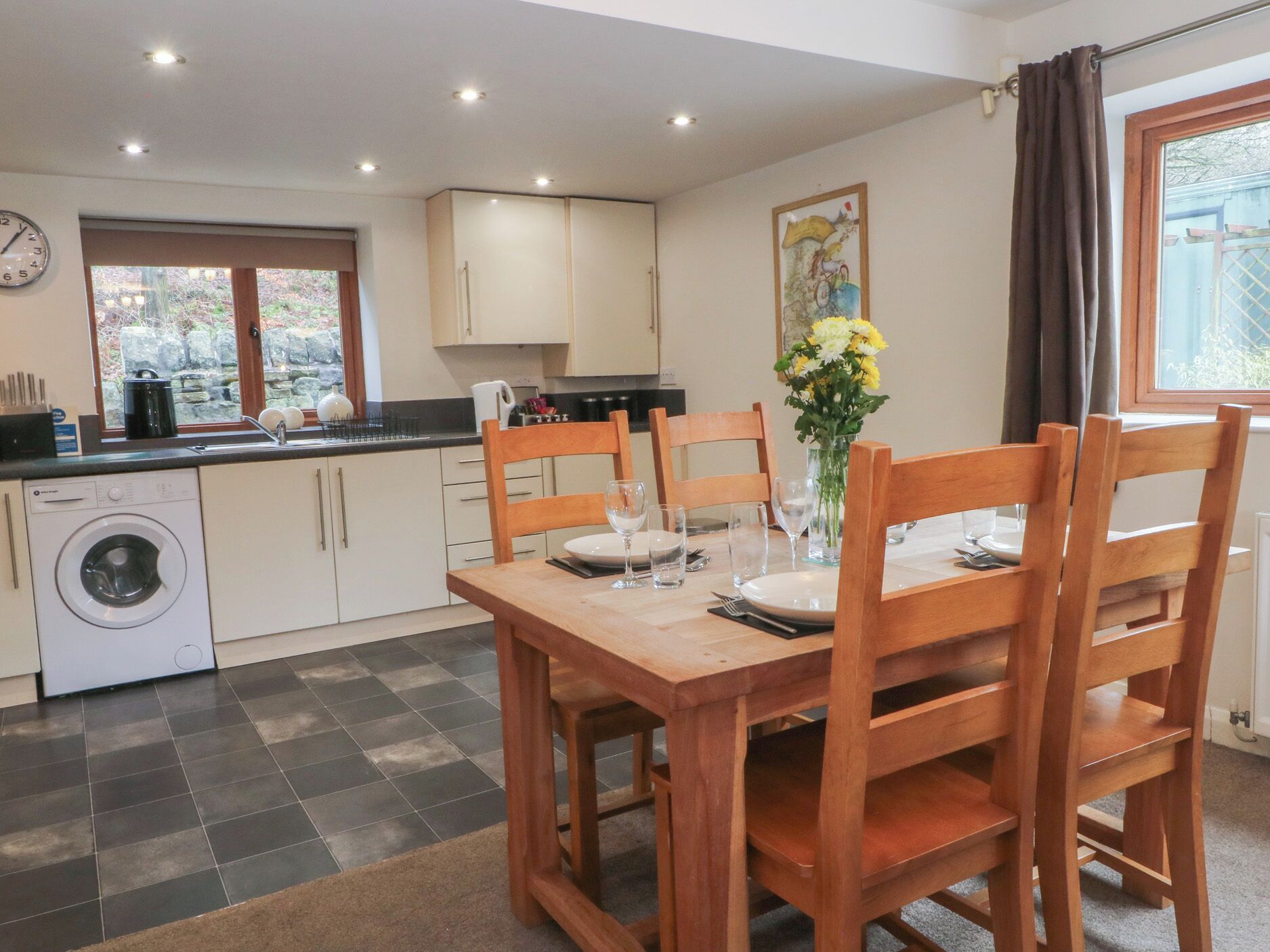 A compact kitchen with a washing machine, a dining table set for four, and a window with curtains.