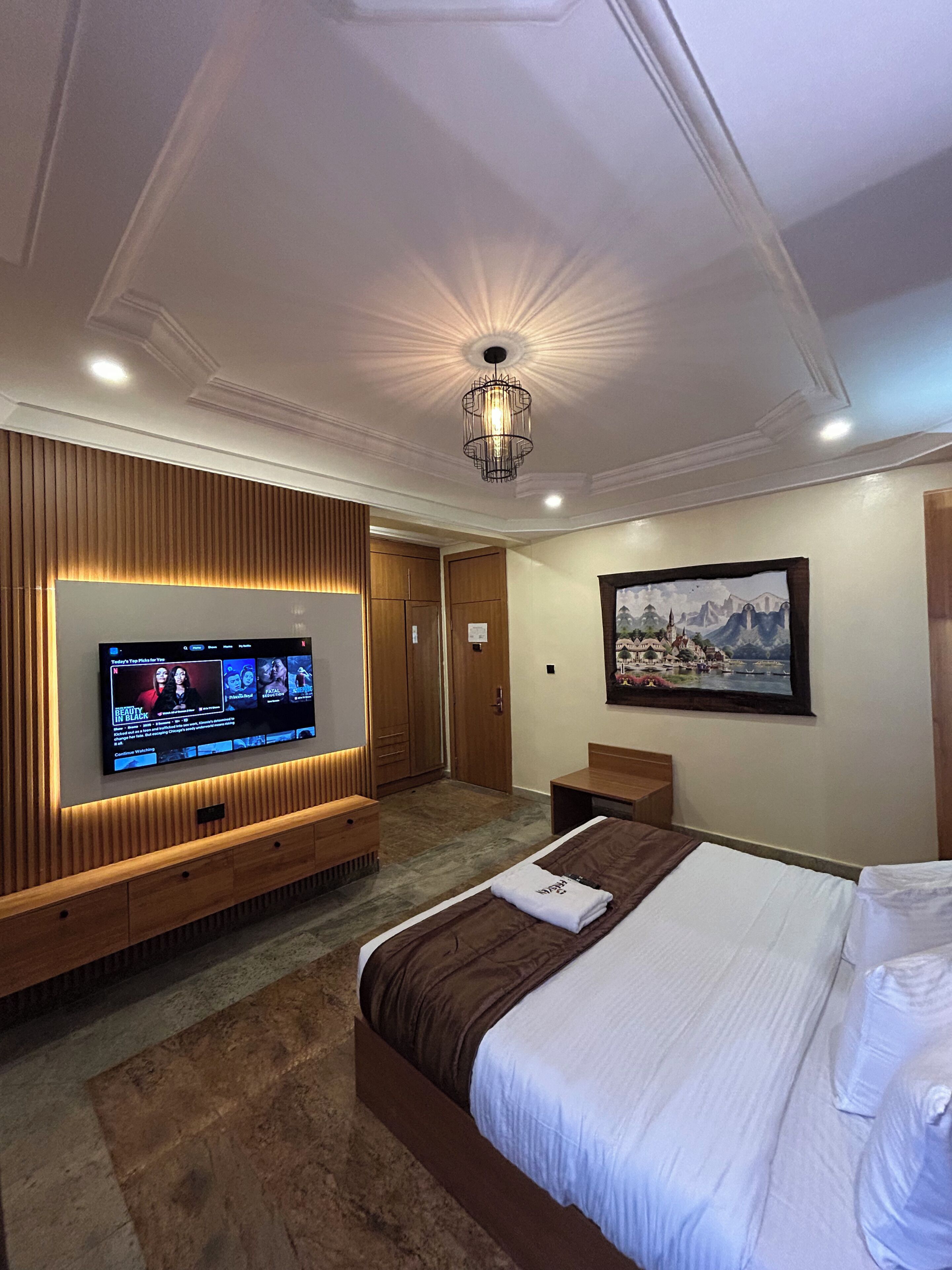 A hotel room with a large TV, a bed, a wooden panel wall, a painting, and a ceiling light fixture.
