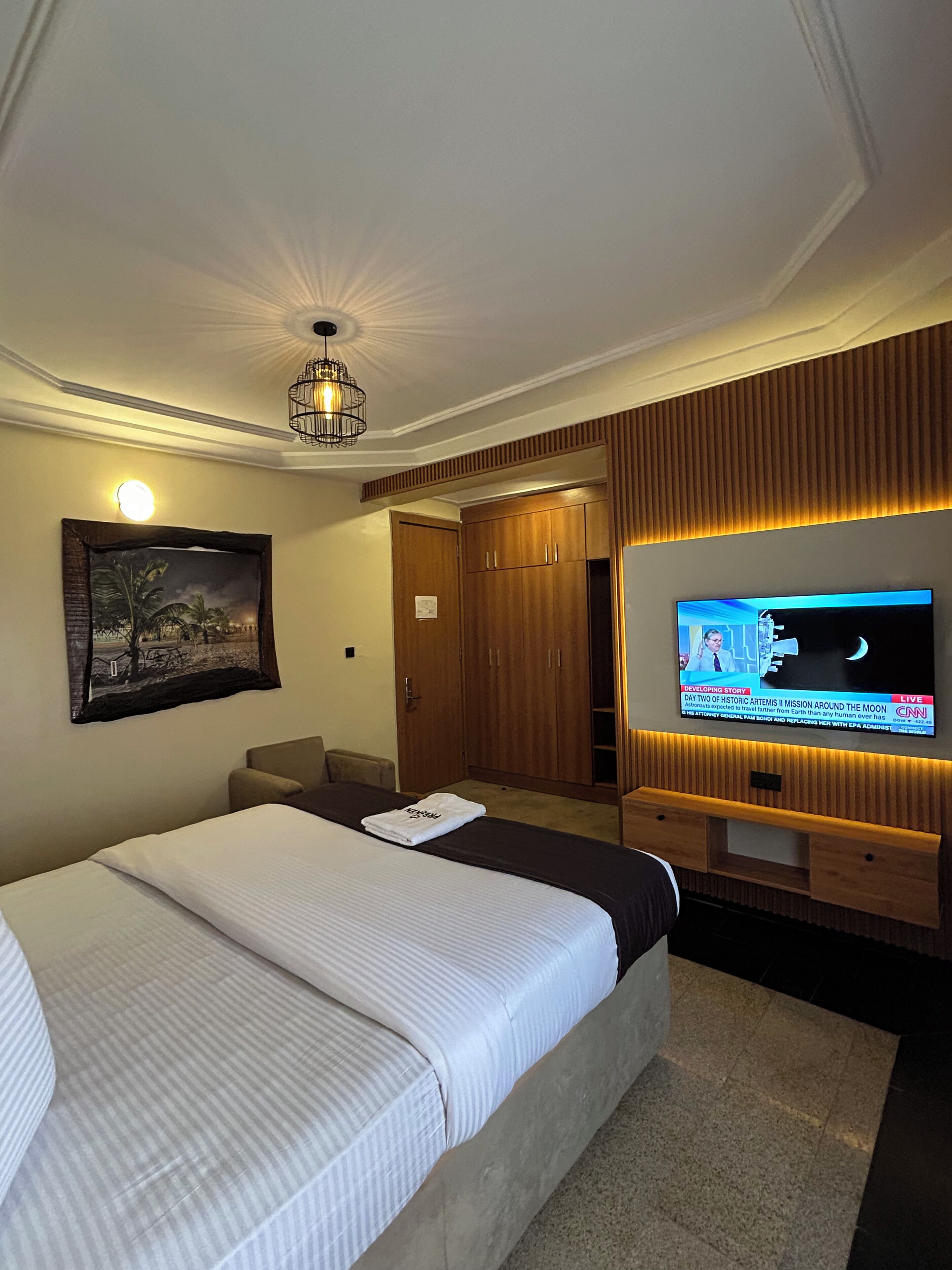 A hotel room with a large bed, a television, a wooden wardrobe, and a painting on the wall.