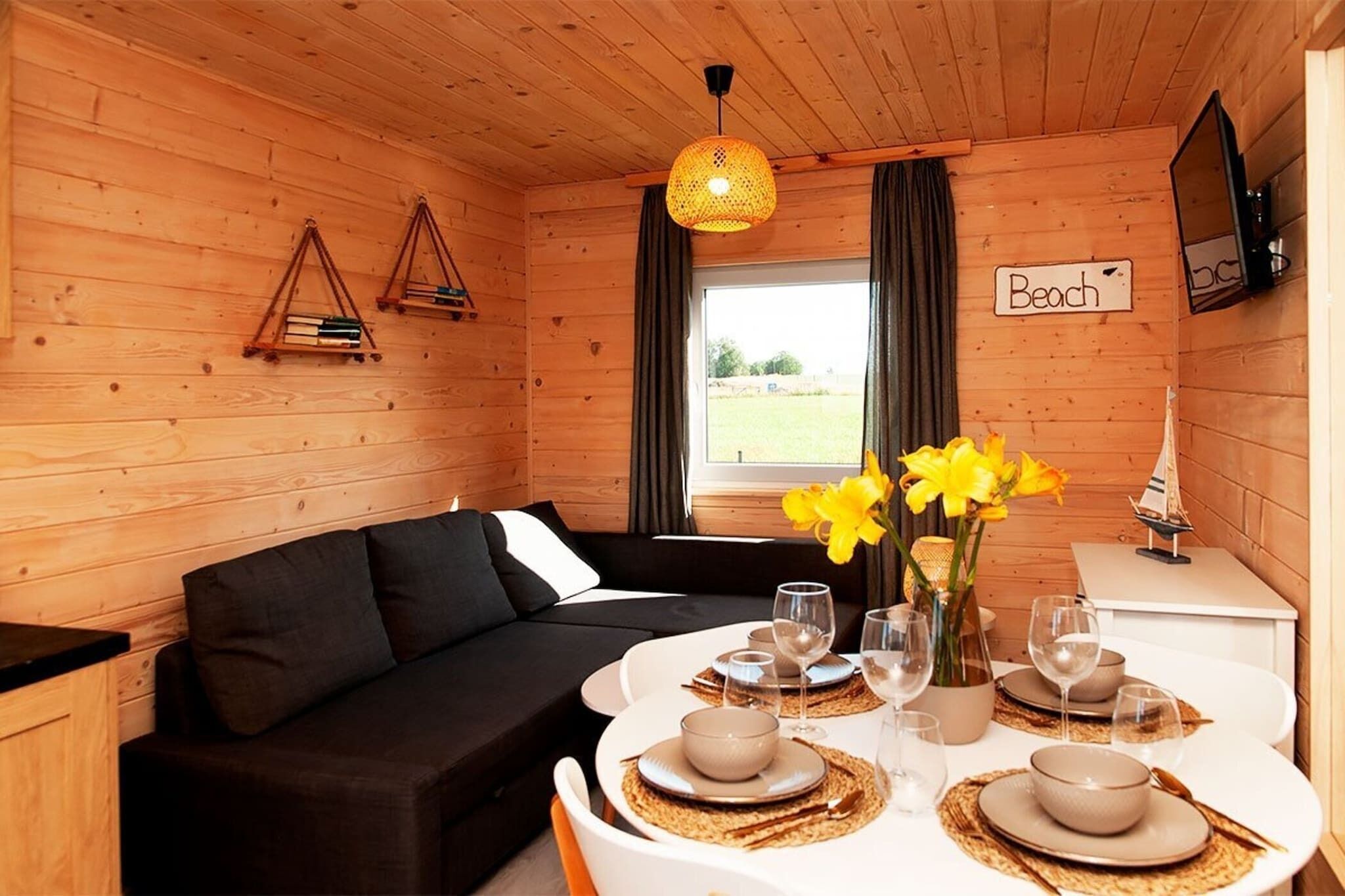Ein gemütliches Blockhaus-Interieur mit einer schwarzen Couch, einem Esstisch für vier Personen und einem Fenster mit Vorhängen.