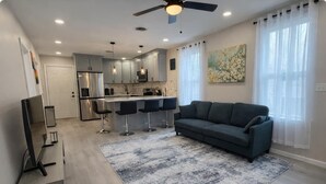 Ein modernes Wohnzimmer mit einem dunklen Sofa, einem Couchtisch und einer TV-Konsole. Eine Küchenzeile mit Kühlschrank, Herd und Barhocker ist ebenfalls zu sehen. Außerdem sind ein Deckenventilator und ein Gemälde sichtbar.