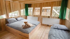 A wooden cabin bedroom with a bed, two smaller beds, a teddy bear, and a window with curtains. - Wooden Lodge Victor de Vos | Slapen in een echte boomhut! (Rijen)