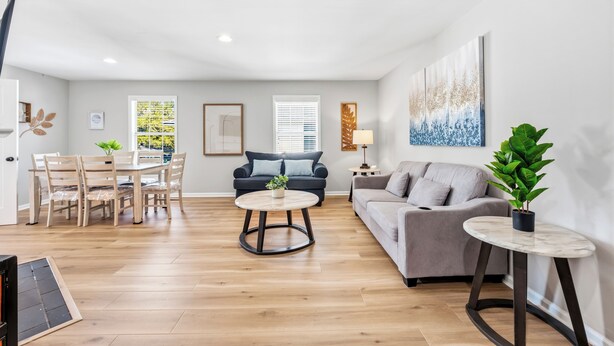 A spacious living room with a wooden floor, a round coffee table, a grey sofa, and a dining area with a table and chairs. - Beach Square 3 –optional BIKES – Walk 2 Redfern Village, shops, and restaurants! (St. Simons Island)