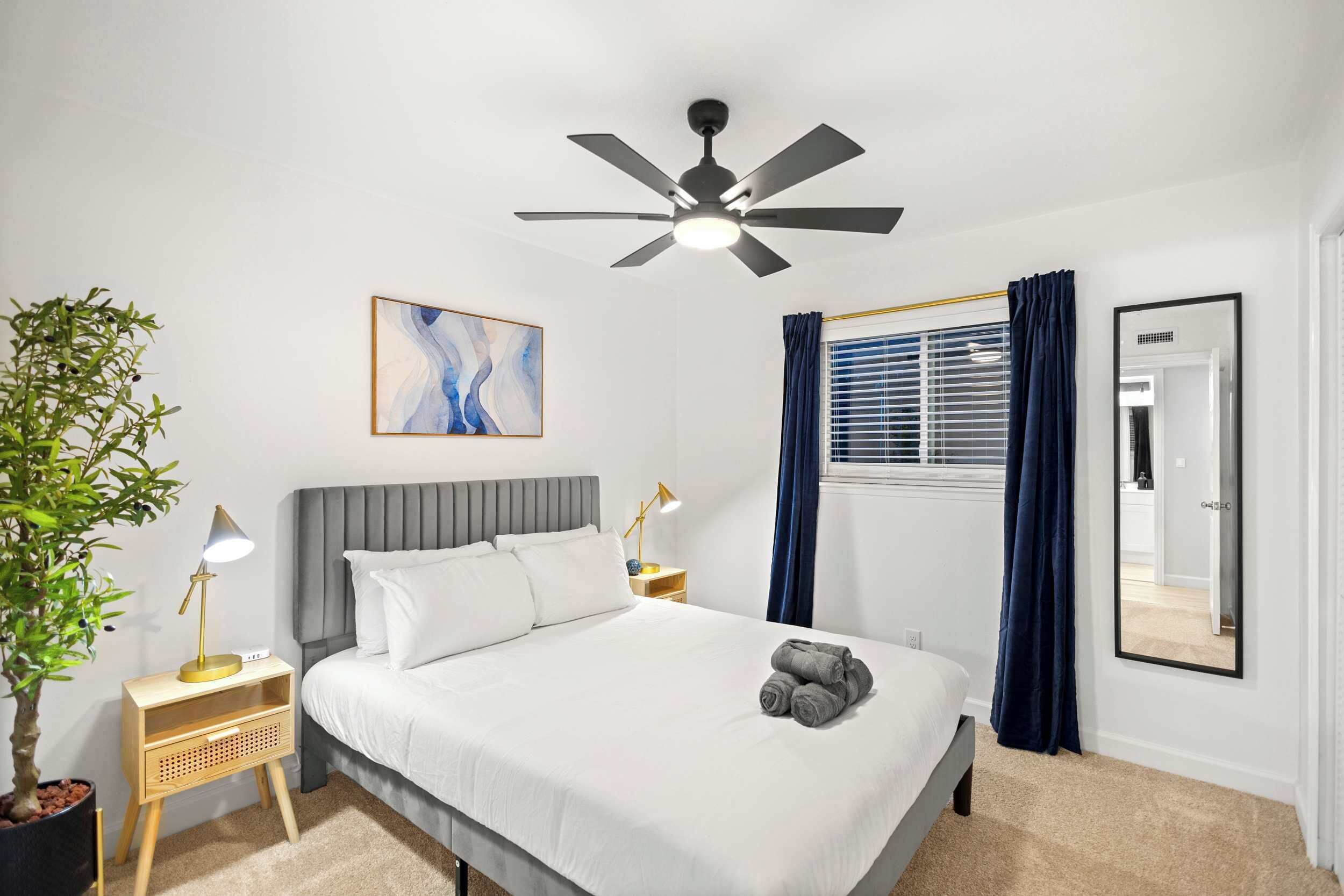 A neatly arranged hotel room with a bed, bedside tables, a ceiling fan, a potted plant, a painting, and a large mirror.