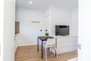 Ein modernes, minimalistisches Zimmer mit Holzboden, weißen Wänden, einem kleinen Esstisch mit Stuhl, einem Fernseher auf einem weißen Schrank und einer kleinen Pflanze auf dem Tisch.