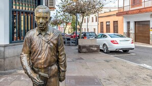 En statue af en mand i jakkesæt, der holder en bog, står på fortov med bygninger og parkerede biler i baggrunden.