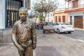 Eine Statue eines Mannes im Anzug, der ein Buch hält, steht auf einem Bürgersteig. Im Hintergrund sind Gebäude und parkende Autos zu sehen.