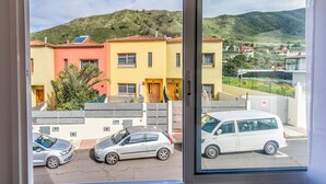 Udsigt fra et vindue over farverige bygninger, en parkeret bil og et kuperet landskab.