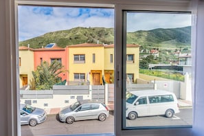 Blick aus einem Fenster auf farbenfrohe Häuser, ein parkendes Auto und eine hügelige Landschaft.