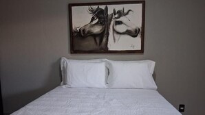 A bed with white bedding and two pillows, a painting of horses on the wall above, and a power outlet on the wall to the right. - Pousada Casa de Naná (Palmas)