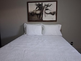 Una cama con ropa blanca y dos almohadas, un cuadro de caballos en la pared superior, y un enchufe en la pared a la derecha.