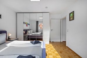 A modern bedroom with a bed, bedside table, lamp, wardrobe, and a view of a cityscape through a window. - Dream on the Old Town Island (Lübeck)