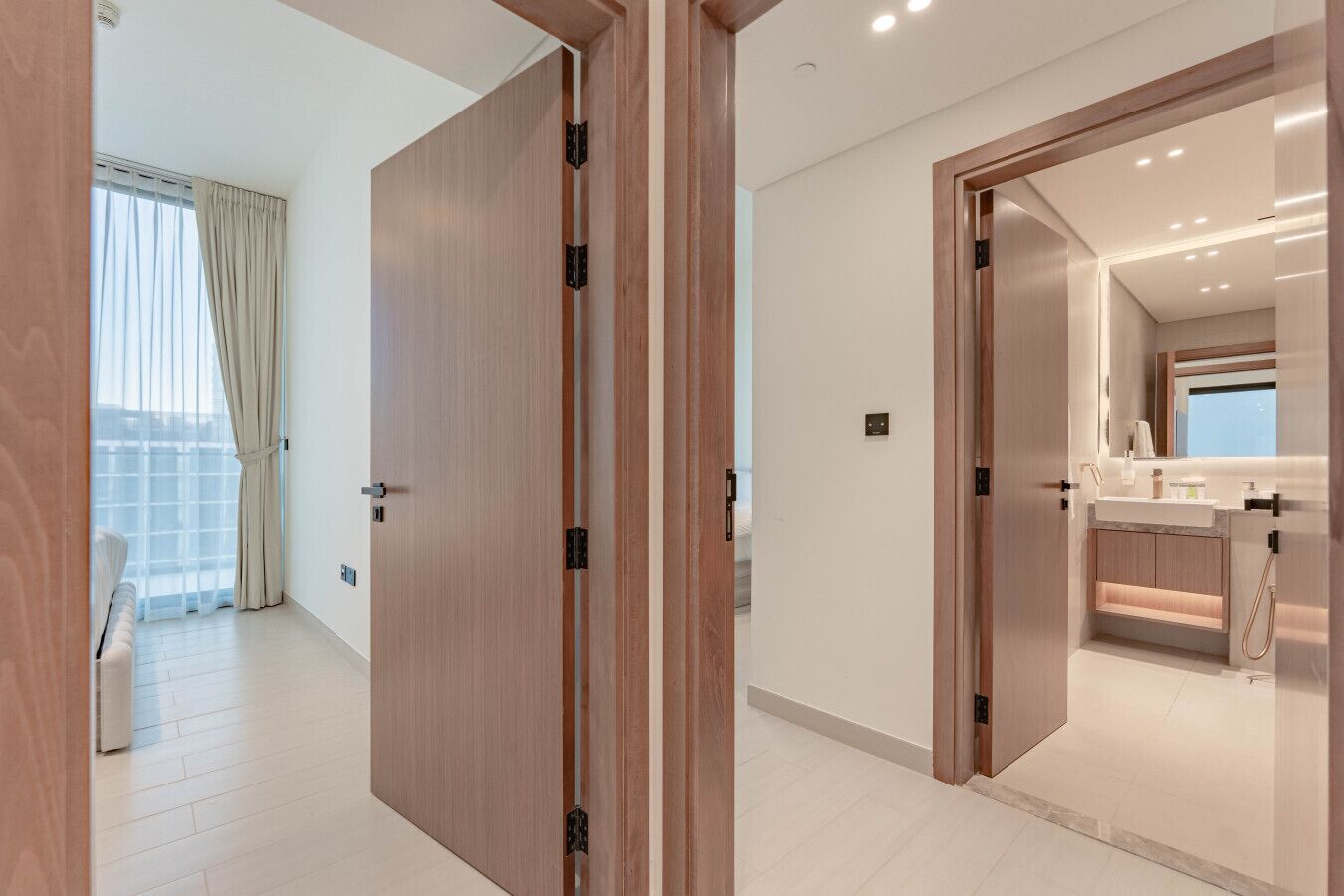 A modern hotel room with a large sliding door, a balcony with curtains, and a well-lit bathroom with a vanity.