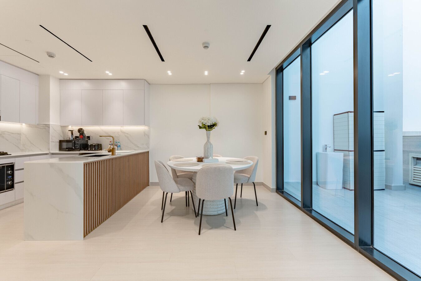 Una cocina moderna con una isla central, una zona de comedor con una mesa redonda y sillas, y amplios ventanales de piso a techo.