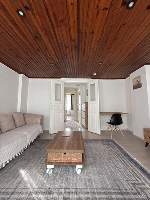 A modern living room with a wooden ceiling, a sofa, a coffee table, and a view into the kitchen. - Stay in the Heart of İstanbul – Modern 1+1 Apartment with Balcony & City View (İstanbul)