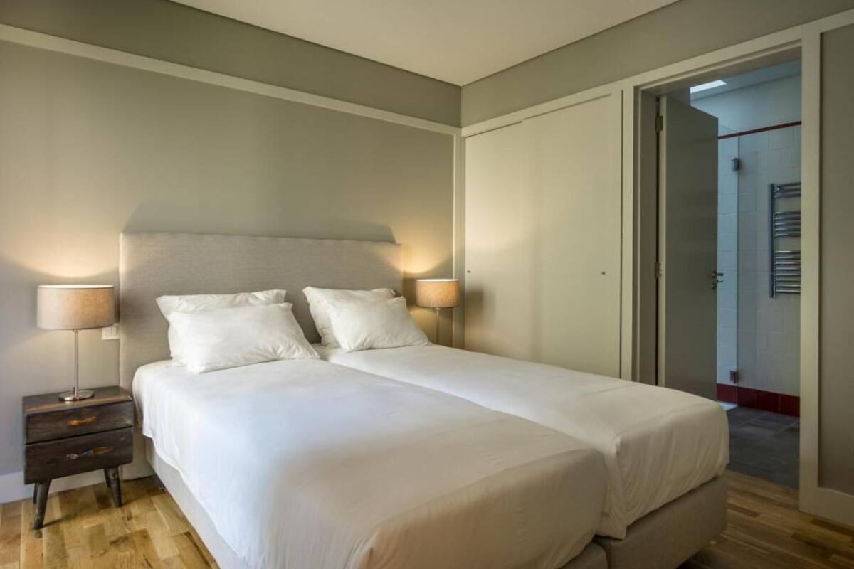Una cama doble con ropa de cama blanca, una mesita de noche con lámpara y una puerta que da a un baño.