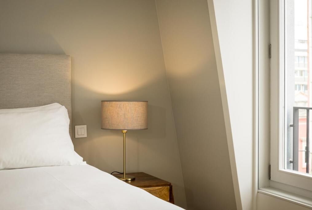 Una camera d'hotel con un letto, un comodino con una lampada e una finestra vista edifici.