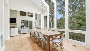 A spacious dining area with a long wooden table and wicker chairs, large windows offering a view of greenery, and a modern kitchen in the background. - Private Marshfront Oasis w/ Pool & Sunset Views (Hilton Head Island)