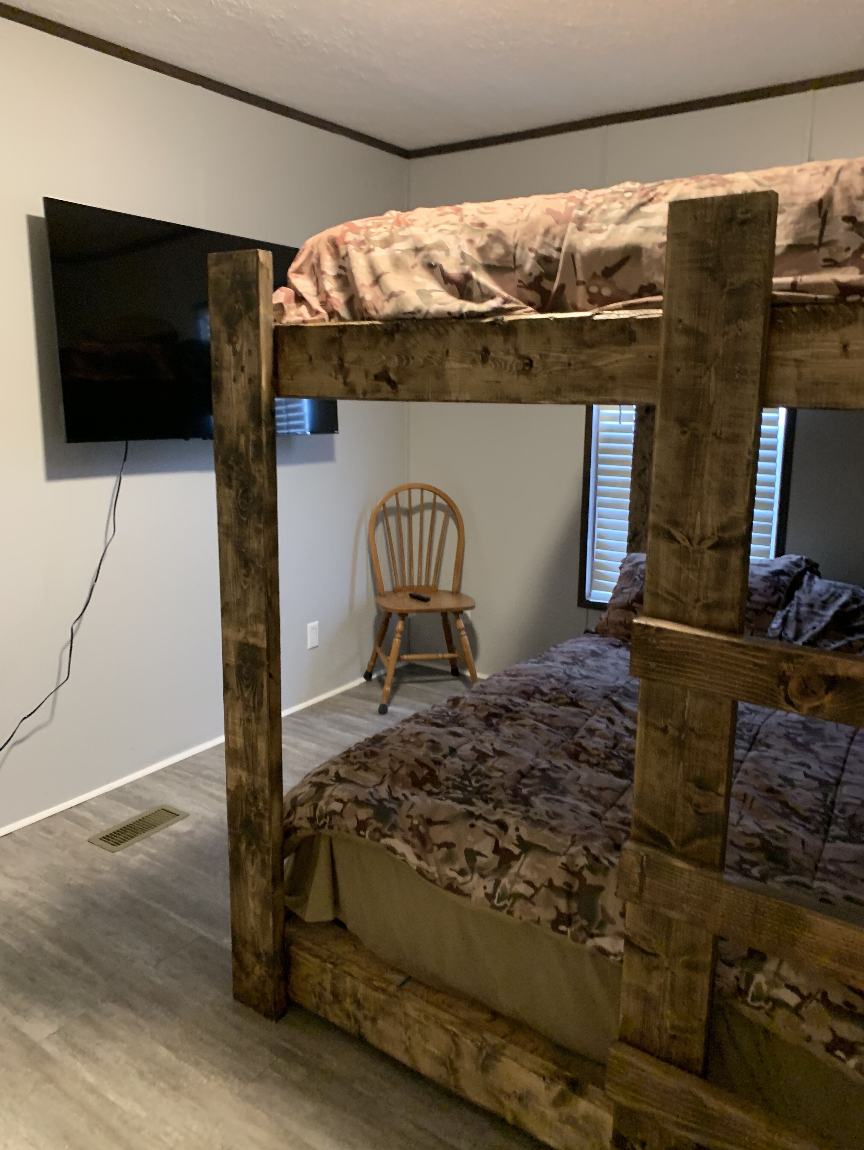 A bunk bed with a camouflage-patterned top and bottom bedding, a wooden chair, and a television mounted on the wall.