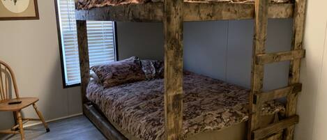 A bunk bed with a camouflage-patterned top sheet, a wooden ladder, and a single pillow. There is a wooden chair and a window with blinds in the room.
