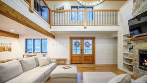 A spacious living room with a high ceiling, a wooden balcony, a fireplace, and a large window. - Sleeps 18 - Hot Tub - Playground - Games - Firepit (Broken Bow)