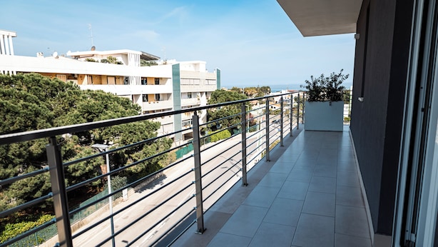 A balcony with a metal railing, a potted plant, and a view of a road and buildings. - D'Stay (Bari)
