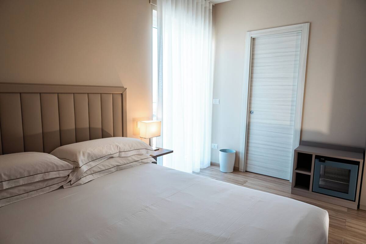 A bedroom with a bed, nightstand, lamp, window with curtains, and a built-in heater.
