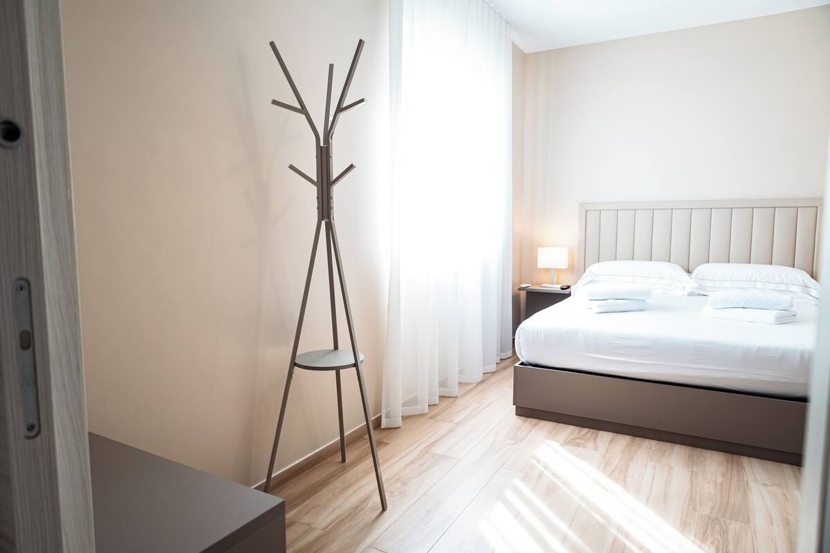 A hotel room with a bed, a nightstand, a lamp, a coat rack, and a wooden floor.
