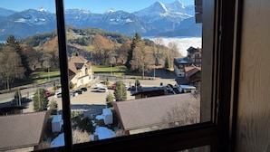 Une vue depuis une fenêtre donnant sur une ville, avec des montagnes enneigées en arrière-plan, un parking où sont garées plusieurs voitures, et un mélange de bâtiments résidentiels et commerciaux.