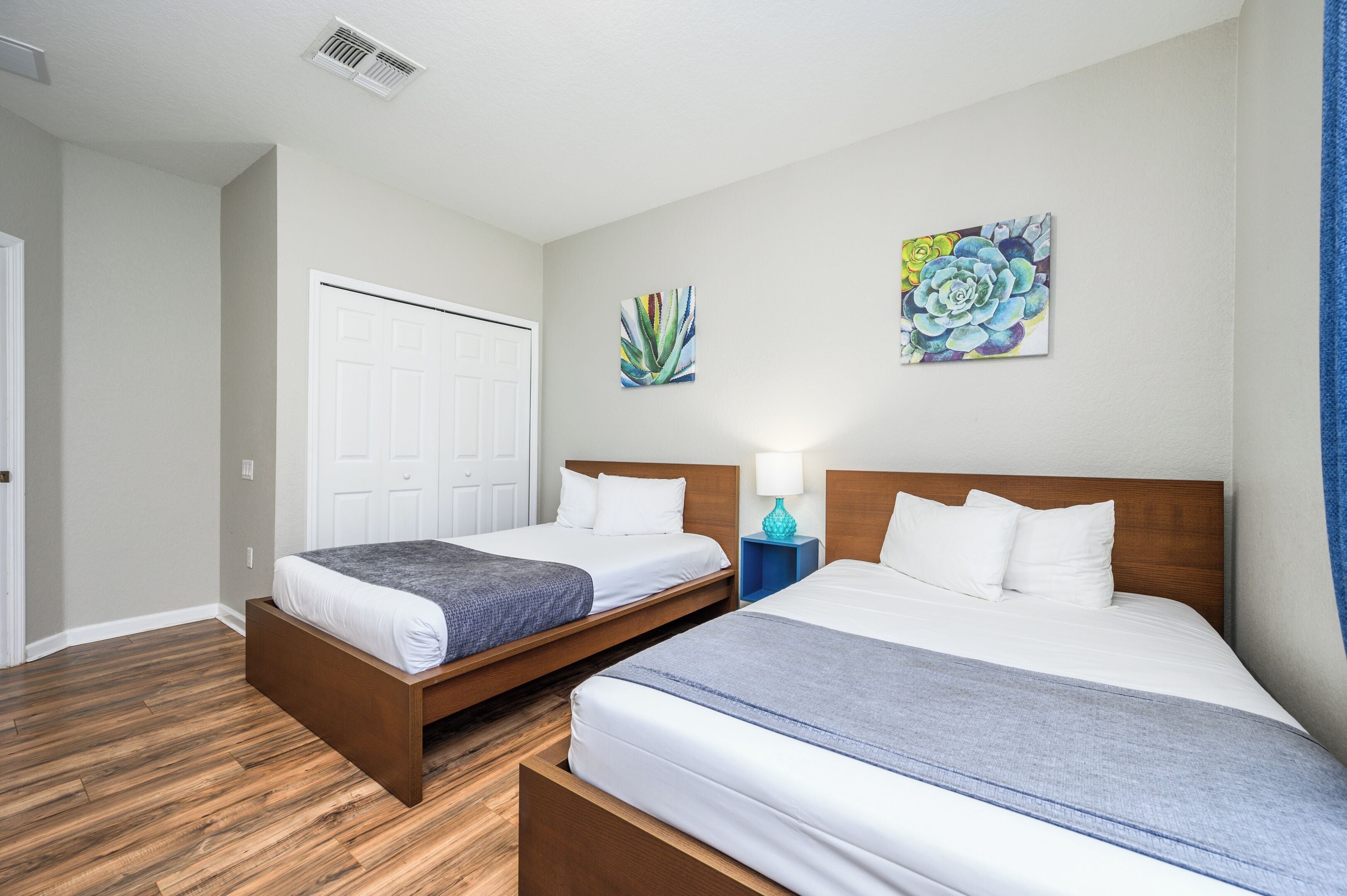 A hotel room with two single beds, a wooden floor, a white door, and two wall-mounted paintings.