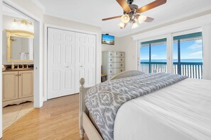 A bedroom with a bed, a ceiling fan, a dresser, a vanity with a mirror, and a view of the ocean through large windows.