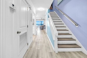 Ein Flur mit einer weißen Tür, einem Lüftungsgitter und einer Treppe mit Holzhäusern und einem Metallgeländer.