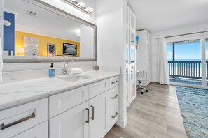 Ein modernes Badezimmer mit großem Spiegel, weißen Schränken, einer Marmorarbeitsplatte und einem Waschbecken. Ein großes Fenster bietet Blick auf den Strand.