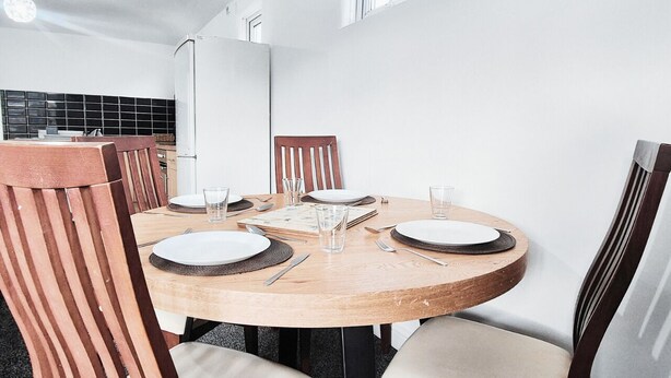 A round wooden dining table with white plates and cutlery, surrounded by wooden chairs with cushioned seats. - Comfy 2BR Flat on Hucknall Road (Nottingham)