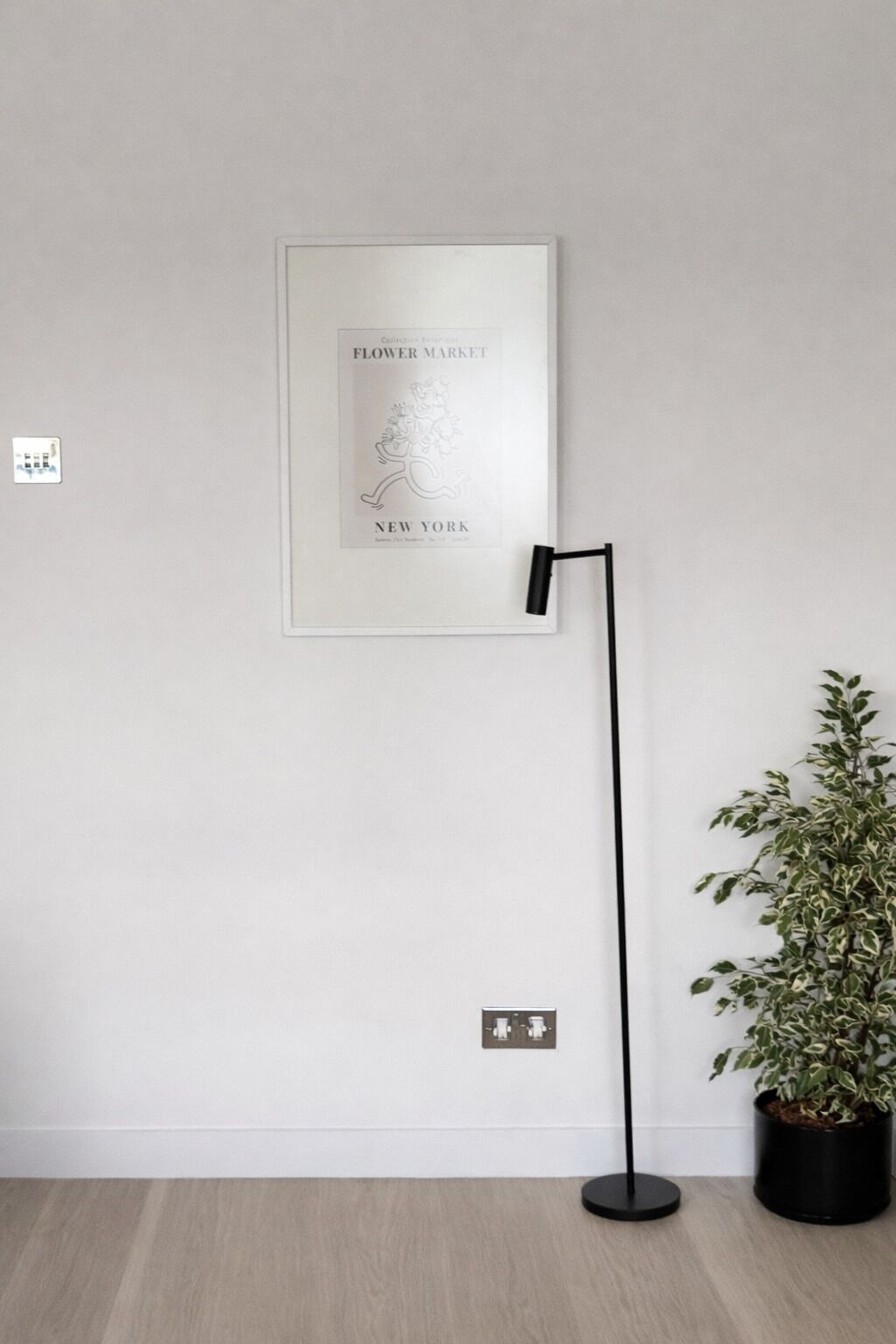 Een lijst met een bloemenmarktadvertentie aan de muur, een vloerlamp en een potplant.