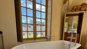 A bathroom with a freestanding bathtub, a large window with a view of a building, and a wooden cabinet with a mirror. - Gite des Cerfs (La Ferté-Saint-Aubin)