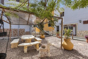 Une terrasse extérieure avec une table ronde et des bancs, entourée de plantes en pot et d’une pergola.