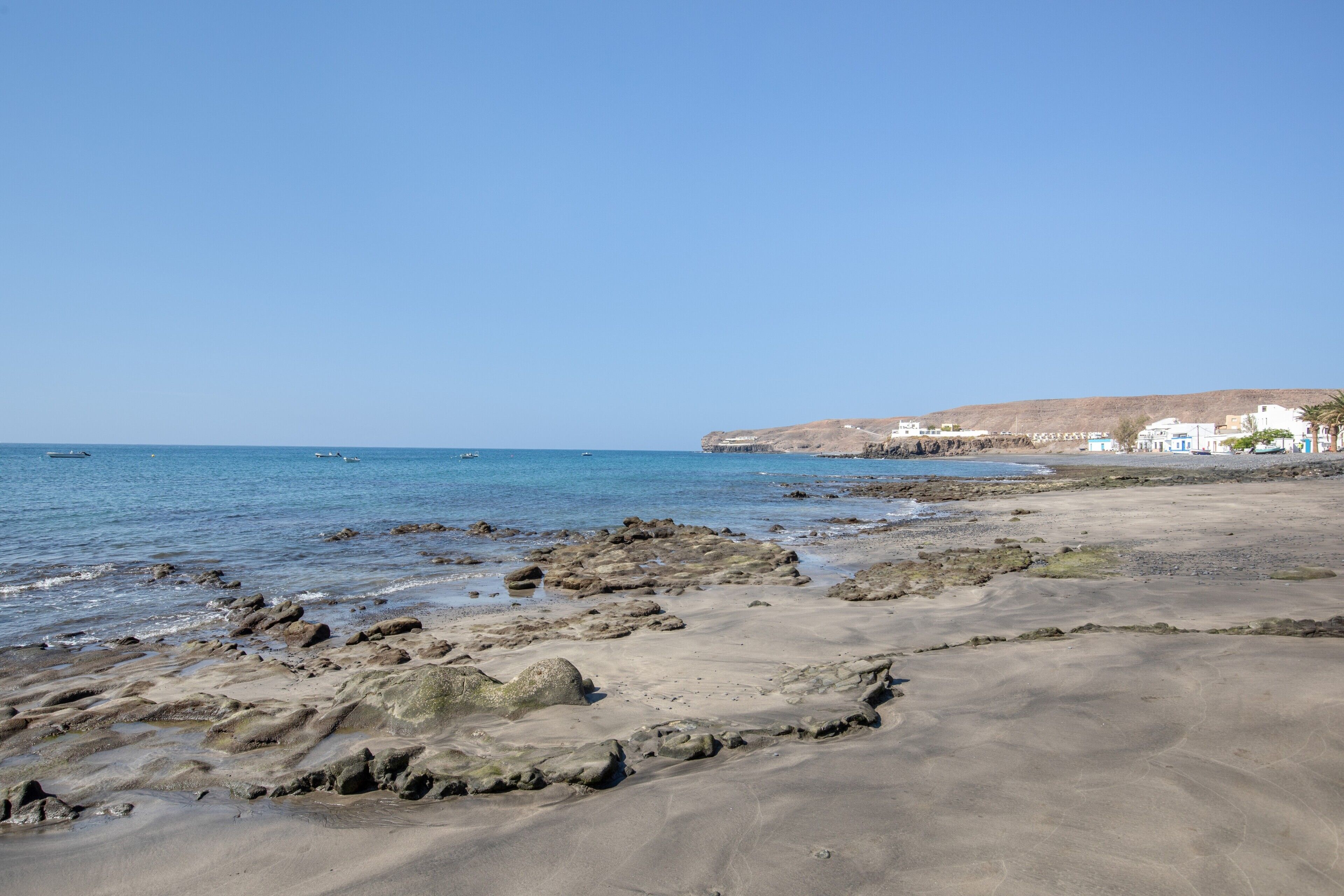 Une plage au paysage rocheux, avec une eau d’un bleu limpide et des bâtiments visibles à l’arrière-plan.