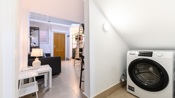 A compact living space with a washing machine, a small table with a lamp, a sofa, and a staircase leading to a loft area. - M Iri70 Acogedor D Plex en el Barrio Salamanca (Madrid)
