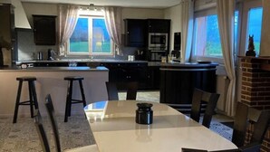 Zona pranzo con un tavolo grande e sedie, cucina sullo sfondo e vista all'esterno attraverso una finestra.
