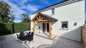 Ein modernes Haus mit überdachter Terrasse, einem Esstisch mit Stühlen und einem kleinen Garten.
