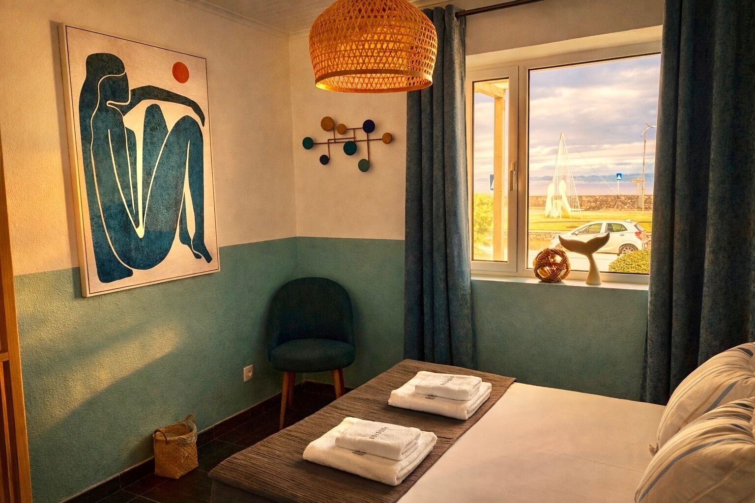 Una camera d'albergo con un letto, una sedia, un tavolo con asciugamani, un quadro, una lampada a sospensione e vista su una barca a vela dalla finestra.