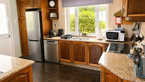 A kitchen with wooden cabinets, a stainless steel refrigerator, a dishwasher, a microwave, and a window with a view of greenery. - Pico Island Villas 117 - Natural Pools and Incredible Views (São Roque do Pico)