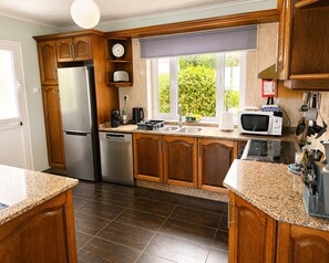A kitchen with wooden cabinets, a stainless steel refrigerator, a dishwasher, a microwave, and a window with a view of greenery. - Pico Island Villas 117 - Natural Pools and Incredible Views (São Roque do Pico)