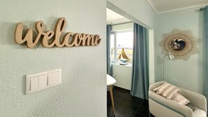 Una stanza con un cartello che riporta la scritta 'Welcome', un interruttore, uno specchio con cornice a forma di sole e un divano con un cuscino.